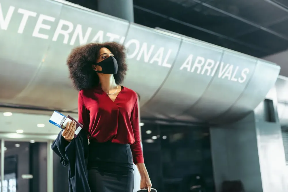 Woman standing at international arrivals terminal holding a boarding pass and wearing a face mask